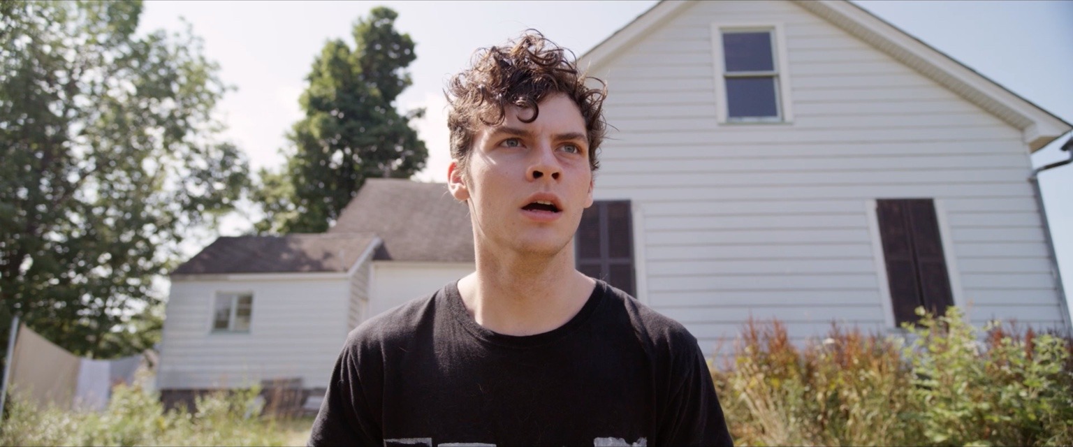 A young man stands outside a white rural house